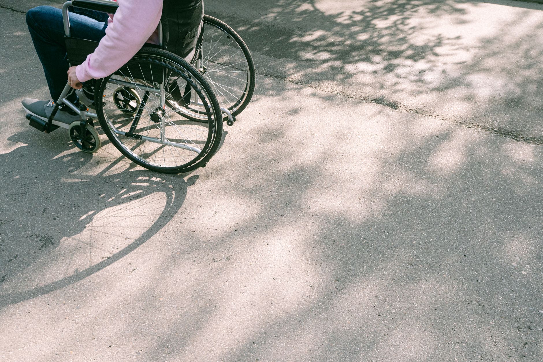 Person using a wheelchair on a sunlit, tree-shaded road.